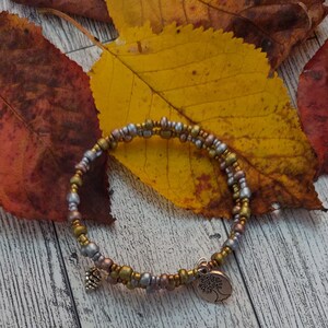 Könnte beinhalten: Ein goldfarbenes, silbernes und kupferfarbenes Perlenarmband mit einem Baum-Anhänger und einem kleinen Tannenzapfen-Anhänger. Das Armband liegt auf einer Holzoberfläche mit Herbstblättern.