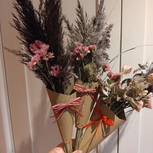May include: Three bouquets of dried flowers in brown paper cones, tied with pink and orange raffia bows. The bouquets feature dark, feathery grasses and pink and beige dried flowers. The bouquets are held in a person's hand.