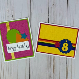 May include: Two handmade birthday cards. One card has a pink background with a green border, a green scalloped circle, and the words "happy birthday". The other card has a yellow background with a red border and the number "8".