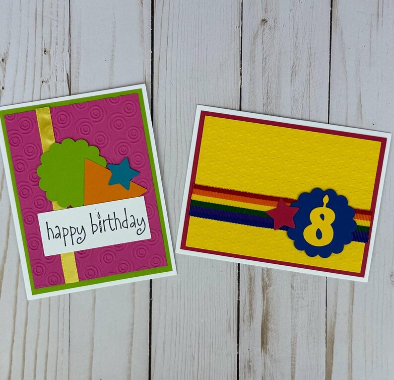 May include: Two handmade birthday cards. One card has a pink background with a green border, a green scalloped circle, and the words "happy birthday". The other card has a yellow background with a red border and the number "8".