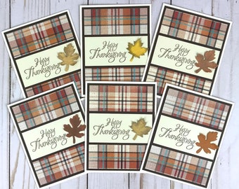 May include: Set of six Thanksgiving greeting cards. Each card features a plaid pattern in shades of orange, brown, and teal. The cards have a cream-colored banner with the words "Happy Thanksgiving" and a leaf embellishment.
