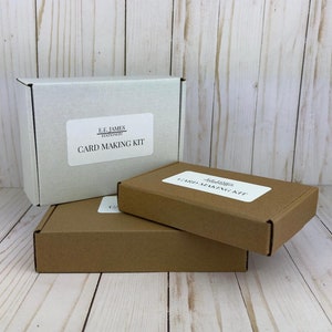 May include: Three rectangular cardboard boxes stacked together. One white box stands upright, while two brown boxes are placed horizontally in front. Each box features a white rectangular label with the text E.F. JAMES STATIONERY CARD MAKING KIT.