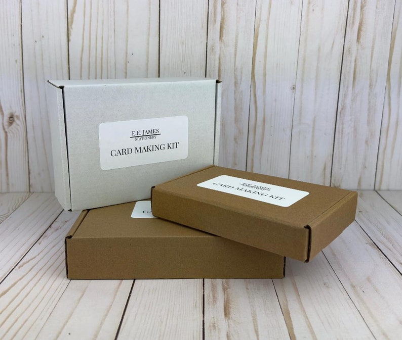 May include: Three rectangular cardboard boxes stacked together. One white box stands upright, while two brown boxes are placed horizontally in front. Each box features a white rectangular label with the text E.F. JAMES STATIONERY CARD MAKING KIT.