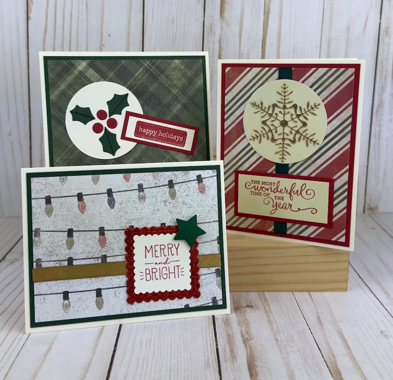Puede incluir: Tres tarjetas de felicitaci&oacute;n rectangulares con motivos festivos. Una tiene un fondo de cuadros verdes con acebo y el texto "happy holidays". Otra tiene rayas rojas y blancas con un copo de nieve dorado y "THE MOST WONDERFUL TIME OF THE YEAR". La tercera muestra luces de cadena y "MERRY and BRIGHT".