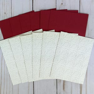 Puede incluir: Un conjunto de tarjetas de felicitaci&oacute;n rectangulares con un panel frontal de color crema con un patr&oacute;n de estrellas geom&eacute;tricas en relieve y un panel posterior de color rojo oscuro s&oacute;lido. Las tarjetas est&aacute;n dispuestas en abanico, mostrando las superficies texturizadas.