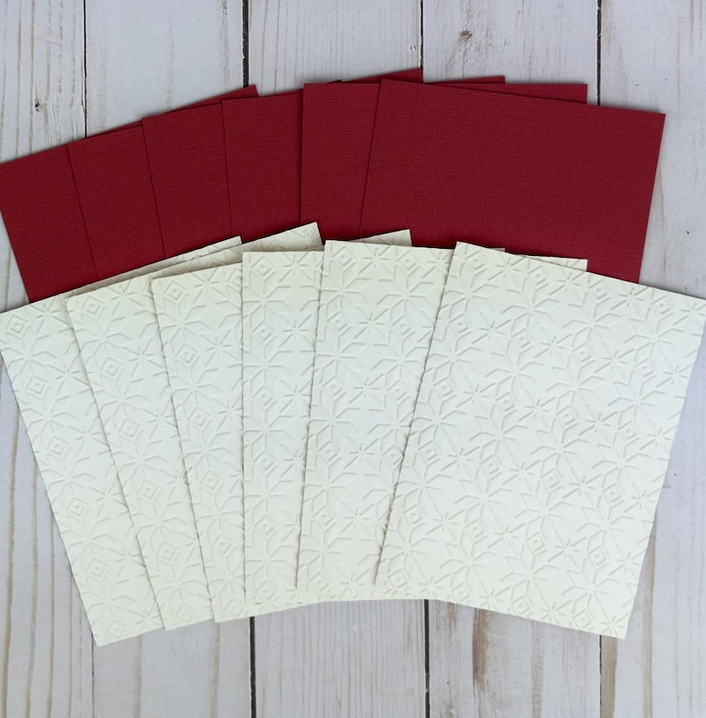 Puede incluir: Un conjunto de tarjetas de felicitaci&oacute;n rectangulares con un panel frontal de color crema con un patr&oacute;n de estrellas geom&eacute;tricas en relieve y un panel posterior de color rojo oscuro s&oacute;lido. Las tarjetas est&aacute;n dispuestas en abanico, mostrando las superficies texturizadas.