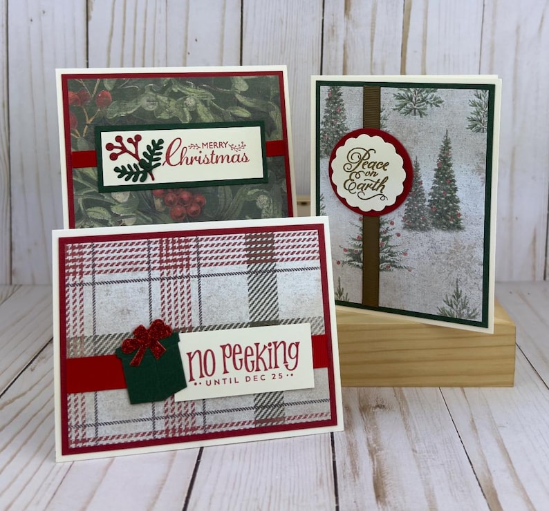 Puede incluir: Tres tarjetas de felicitaci&oacute;n rectangulares con temas navide&ntilde;os. Una tarjeta muestra un patr&oacute;n de acebo con el texto Merry Christmas. Otra muestra un bosque nevado con el texto Peace on Earth. La tercera presenta un patr&oacute;n de cuadros con un icono de regalo y el texto no Peeking UNTIL DEC 25.