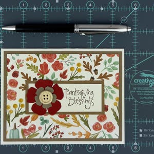 Pu&ograve; includere: Un biglietto d'auguri rettangolare con un motivo di foglie autunnali, fiori e ghiande nei toni del rosso, arancione, giallo e verde. Un'etichetta centrale color crema riporta la scritta "Thanksgiving Blessings" con un fiore di carta rosso e grigio e un bottone in legno.