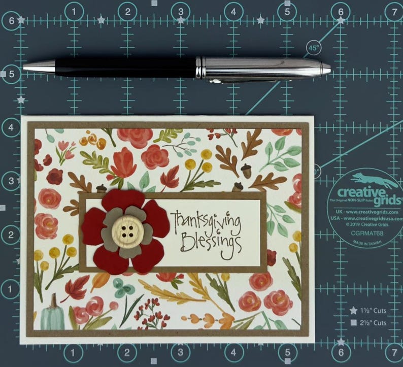 Pu&ograve; includere: Un biglietto d'auguri rettangolare con un motivo di foglie autunnali, fiori e ghiande nei toni del rosso, arancione, giallo e verde. Un'etichetta centrale color crema riporta la scritta "Thanksgiving Blessings" con un fiore di carta rosso e grigio e un bottone in legno.