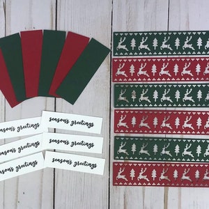 以下が含まれることがあります： 赤と緑のカードストックの長方形のストリップ6枚、黒い文字でseason's greetingsと印刷された白い長方形のラベル6枚、トナカイと木の模様が繰り返されるダイカットストリップ6枚を含むクラフト用品のセット。
