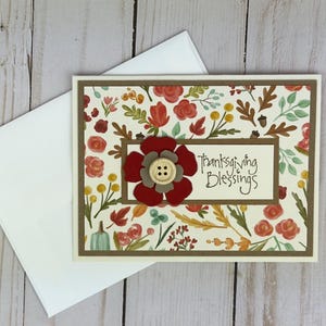 Pu&ograve; includere: Un biglietto d'auguri rettangolare con sfondo color crema caratterizzato da un motivo di fiori rossi, foglie arancioni, bacche gialle e piccole ghiande. Un pannello centrale mostra il testo Thanksgiving Blessings con un fiore rosso e un bottone in legno.