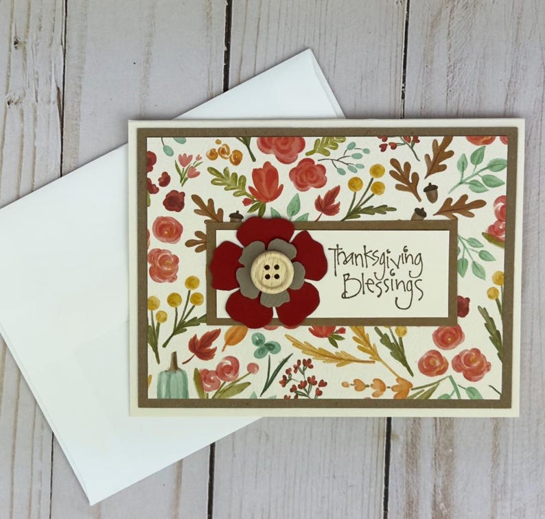Pu&ograve; includere: Un biglietto d'auguri rettangolare con sfondo color crema caratterizzato da un motivo di fiori rossi, foglie arancioni, bacche gialle e piccole ghiande. Un pannello centrale mostra il testo Thanksgiving Blessings con un fiore rosso e un bottone in legno.