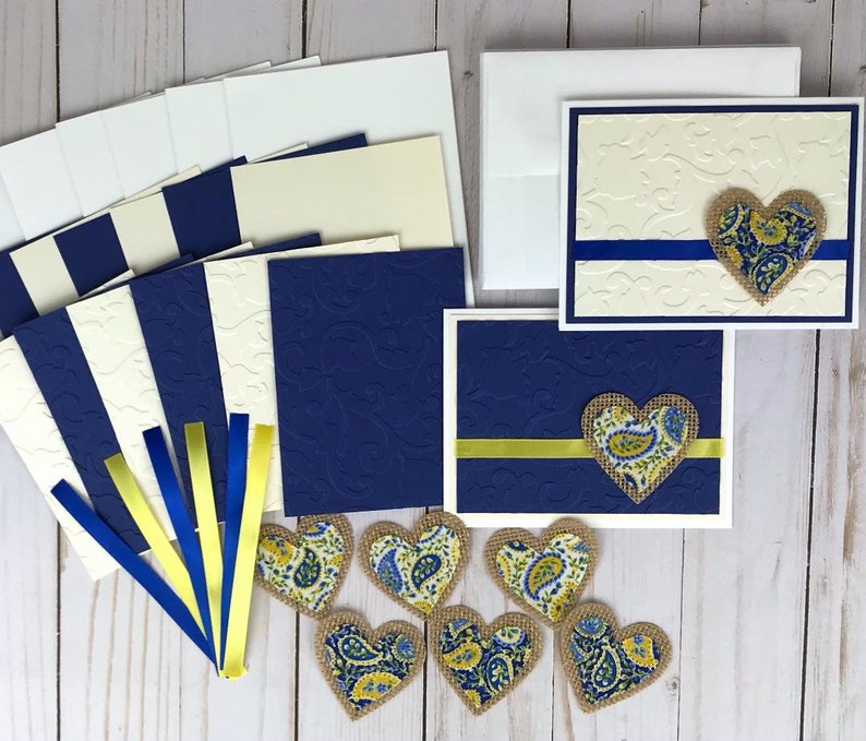 May include: A collection of handmade greeting cards and embellishments. The cards feature embossed designs and are accented with blue and yellow ribbons and paisley heart-shaped appliqu&eacute;s. Includes envelopes and blank cardstock.