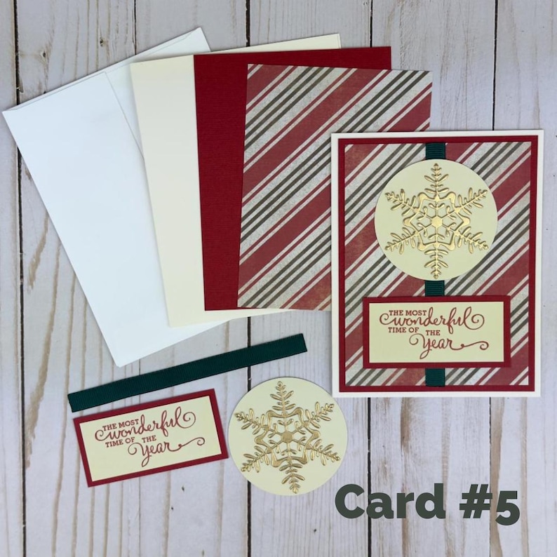 Puede incluir: Un juego de tarjetas de felicitaci&oacute;n con un patr&oacute;n de rayas rojas y blancas, un c&iacute;rculo color crema con un copo de nieve dorado y una etiqueta rectangular con el texto "The most wonderful time of the year" en letra roja, junto a un sobre blanco.