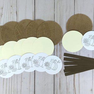 May include: A collection of craft supplies including several tan burlap circles, pale yellow cardstock circles, white cardstock circles featuring a black line art illustration of a pumpkin and sunflower, and six strips of dark brown ribbon.