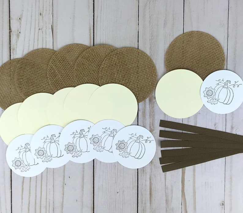 May include: A collection of craft supplies including several tan burlap circles, pale yellow cardstock circles, white cardstock circles featuring a black line art illustration of a pumpkin and sunflower, and six strips of dark brown ribbon.
