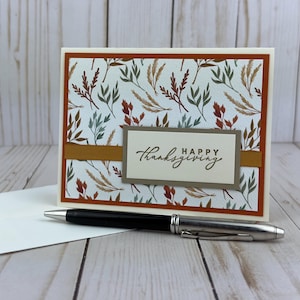 May include: A rectangular greeting card featuring a pattern of autumn-colored leaves in shades of rust, sage green, and tan on a white background. A horizontal mustard yellow ribbon crosses the center, topped with a label reading HAPPY thanksgiving.