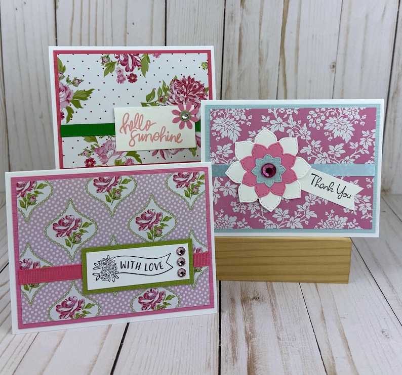 K&ouml;nnte beinhalten: Drei rechteckige Gru&szlig;karten mit Blumenmustern. Eine Karte hat einen wei&szlig;en Hintergrund mit Punkten und rosa Rosen sowie den Text hello sunshine. Eine weitere hat einen rosa Blumendruck mit einer geschichteten Blume und den Text Thank You. Eine hat ein violettes Rosenmuster mit dem Text WITH LOVE.