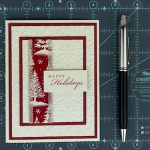 Puede incluir: Una tarjeta de felicitaci&oacute;n rectangular con un patr&oacute;n geom&eacute;trico en relieve de color crema, un borde rojo y una tira de tela roja vertical con &aacute;rboles y copos de nieve blancos. Incluye una etiqueta blanca con el texto "HAPPY Holidays". Un bol&iacute;grafo sirve de escala.
