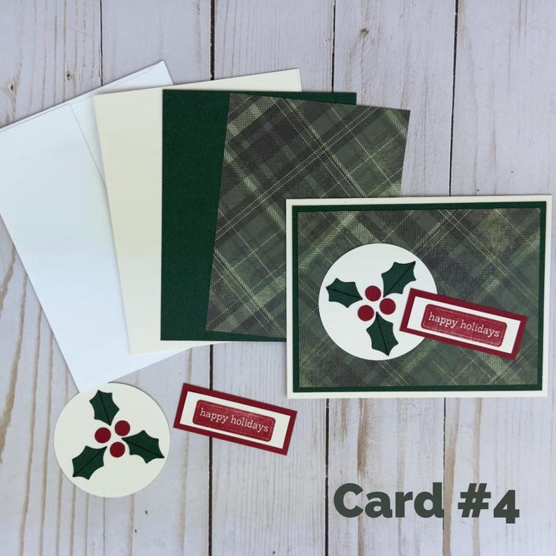 Puede incluir: Un juego de tarjetas de felicitaci&oacute;n con un patr&oacute;n de cuadros verde oscuro, un recorte circular blanco con hojas de acebo verdes y bayas rojas, y una etiqueta rectangular con el texto "happy holidays" en rojo. Incluye sobres y cartulina.