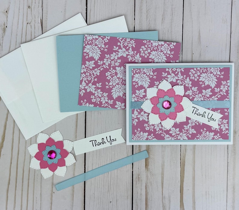 K&ouml;nnte beinhalten: Eine Gru&szlig;karte mit rosa-wei&szlig;em Blumenmuster, einer mehrlagigen Papierblumenverzierung, einem rosa Strassstein in der Mitte und einem wei&szlig;en Banner mit dem Text Thank You. Enth&auml;lt passende Umschl&auml;ge und zus&auml;tzliche Papierbastelkomponenten.