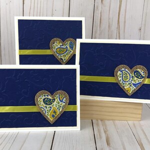 May include: Three handmade greeting cards with a navy blue embossed background and a white border. Each card features a burlap heart with a paisley pattern and a yellow ribbon. The cards are displayed on a light wooden surface.