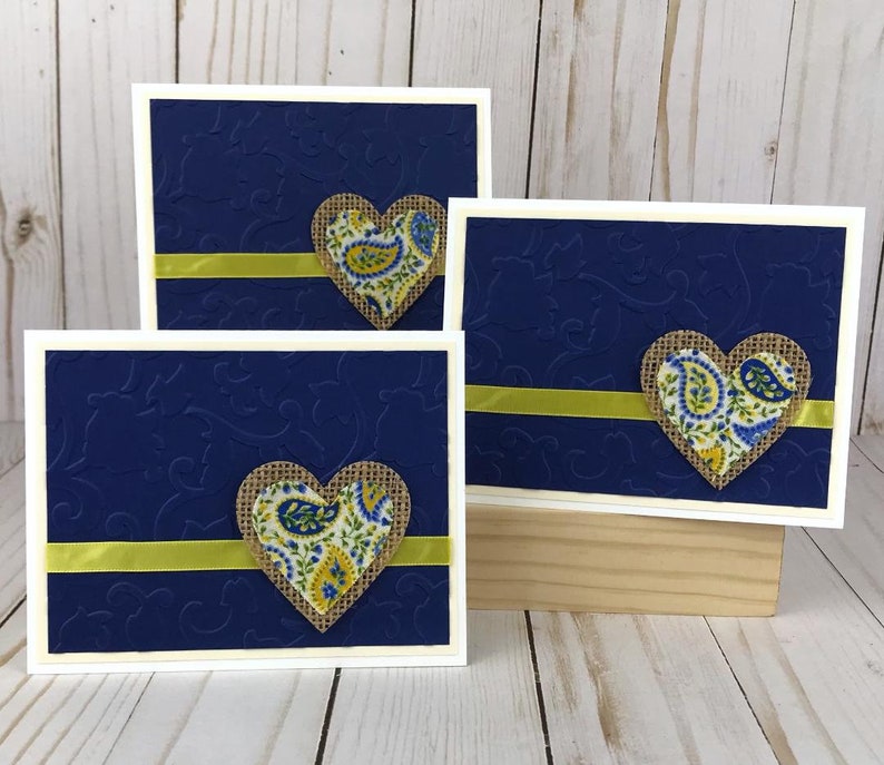 May include: Three handmade greeting cards with a navy blue embossed background and a white border. Each card features a burlap heart with a paisley pattern and a yellow ribbon. The cards are displayed on a light wooden surface.