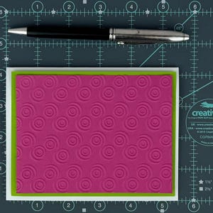 May include: A magenta card with a spiral pattern, framed by a green border and a white border. A black and silver pen rests above the card on a grid background.