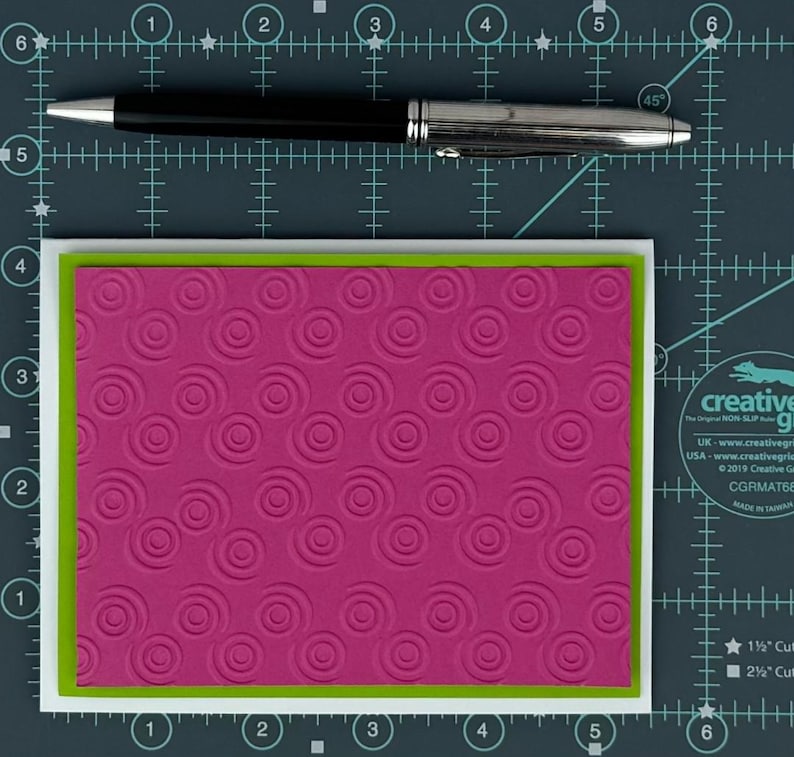 May include: A magenta card with a spiral pattern, framed by a green border and a white border. A black and silver pen rests above the card on a grid background.