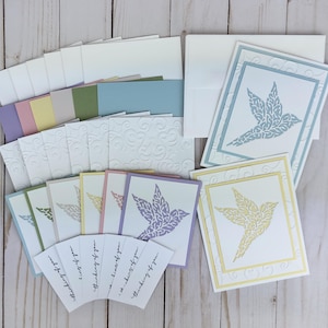May include: A collection of handmade greeting cards with a dove design. The cards are in various pastel colors, including blue, yellow, and lavender. The set includes matching envelopes and cards with the text "thinking of you".
