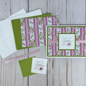K&ouml;nnte beinhalten: Eine Gru&szlig;karte mit einem rosa-wei&szlig; gestreiften Hintergrund mit Blumenmustern, einem horizontalen rosa Band und einem wei&szlig;en quadratischen Etikett mit dem Text "just a little note" und einer kleinen G&auml;nsebl&uuml;mchen-Illustration mit rosa Schmuckstein.