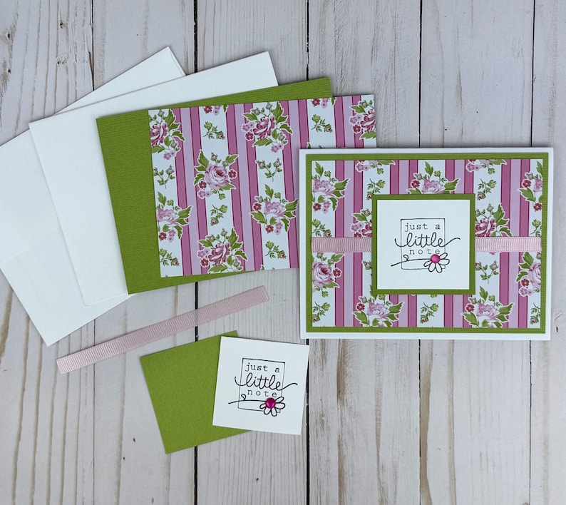 K&ouml;nnte beinhalten: Eine Gru&szlig;karte mit einem rosa-wei&szlig; gestreiften Hintergrund mit Blumenmustern, einem horizontalen rosa Band und einem wei&szlig;en quadratischen Etikett mit dem Text "just a little note" und einer kleinen G&auml;nsebl&uuml;mchen-Illustration mit rosa Schmuckstein.