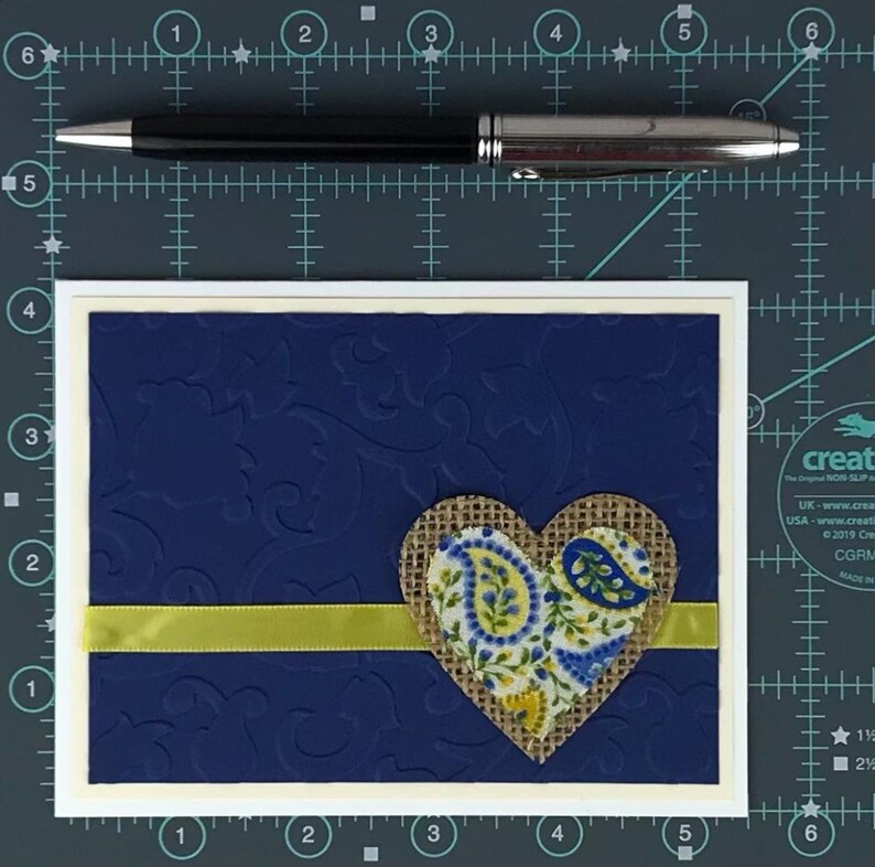 May include: A greeting card with a navy blue embossed background and a white border. A burlap heart with a paisley design is centered on the card, with a yellow ribbon and a black pen.