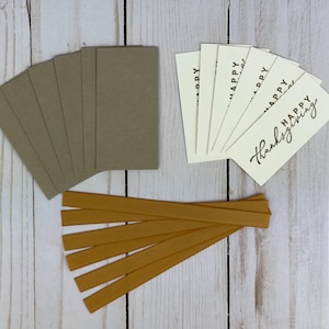 May include: A set of rectangular tags including four textured taupe cards and four cream-colored cards printed with the black text Happy Thanksgiving, accompanied by five strips of mustard yellow ribbed ribbon for attachment and display.