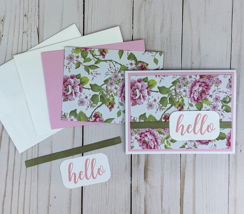 K&ouml;nnte beinhalten: Eine Gru&szlig;karte mit einem Blumenmuster aus rosa Rosen und gr&uuml;nen Bl&auml;ttern auf wei&szlig;em Grund. Ein horizontaler olivgr&uuml;ner Bandstreifen verl&auml;uft durch die Mitte, belegt mit einem wei&szlig;en rechteckigen Etikett, das das Wort hello in Rosa zeigt.