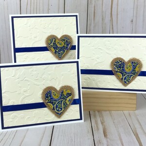 May include: Three cream-colored greeting cards with a white border and a navy blue accent. Each card features a burlap heart with a blue and yellow paisley pattern. The cards are arranged on a wooden surface.