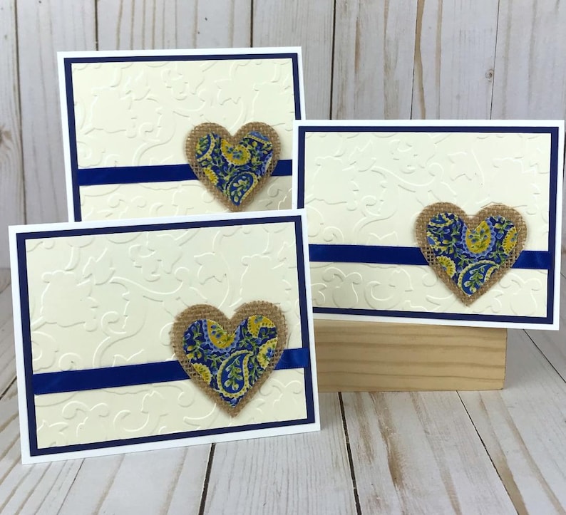 May include: Three cream-colored greeting cards with a white border and a navy blue accent. Each card features a burlap heart with a blue and yellow paisley pattern. The cards are arranged on a wooden surface.