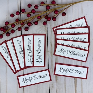 Puede incluir: Una colección de etiquetas de papel rectangulares con fondo blanco, borde rojo oscuro y el texto Merry Christmas escrito en letra cursiva negra. Una rama decorativa con pequeñas bayas de color rojo oscuro se sitúa sobre las etiquetas.