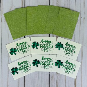 May include: A collection of St. Patrick's Day cards and glitter paper. The cards are off-white with a green shamrock and the text "Happy St. Patrick's Day." The glitter paper is a bright green color.