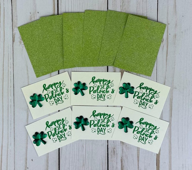 May include: A collection of St. Patrick's Day cards and glitter paper. The cards are off-white with a green shamrock and the text "Happy St. Patrick's Day." The glitter paper is a bright green color.