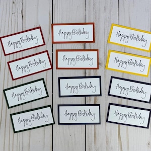 May include: A collection of eleven rectangular paper tags, each featuring the handwritten text Happy Birthday in black ink on a white background. The tags are framed by colored borders in shades of red, orange, yellow, green, and dark blue.