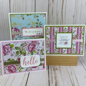 K&ouml;nnte beinhalten: Drei rechteckige Gru&szlig;karten mit Blumen- und Streifenmustern in Rosa-, Gr&uuml;n- und Blaut&ouml;nen. Die Karten zeigen den Text "thinking of you", "just a little note" und "hello" auf geschichteten Papierakzenten mit B&auml;ndern und kleinen Schmucksteinen.