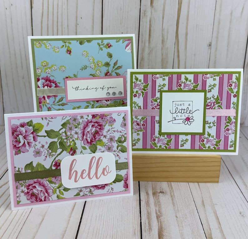 K&ouml;nnte beinhalten: Drei rechteckige Gru&szlig;karten mit Blumen- und Streifenmustern in Rosa-, Gr&uuml;n- und Blaut&ouml;nen. Die Karten zeigen den Text "thinking of you", "just a little note" und "hello" auf geschichteten Papierakzenten mit B&auml;ndern und kleinen Schmucksteinen.