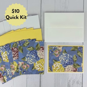 May include: A stationery set featuring rectangular cards with a blue floral pattern of hydrangeas in shades of blue, yellow, and pink. The set includes matching yellow cardstock inserts, plain white envelopes, and a yellow starburst label with text $10 Quick Kit.