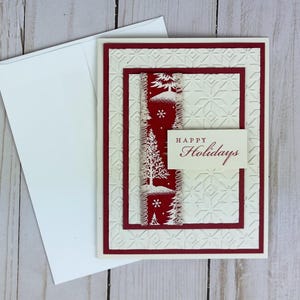 Puede incluir: Una tarjeta de felicitaci&oacute;n de color crema con un patr&oacute;n geom&eacute;trico en relieve, que presenta una franja vertical roja con ilustraciones blancas de pinos y copos de nieve, y una peque&ntilde;a etiqueta rectangular con el texto Happy Holidays.