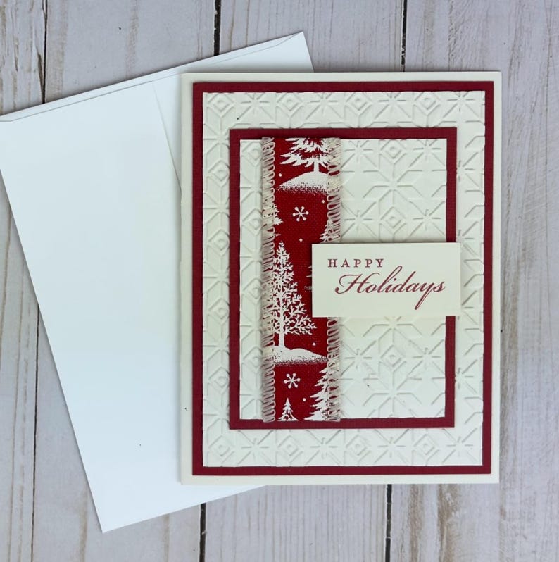 Puede incluir: Una tarjeta de felicitaci&oacute;n de color crema con un patr&oacute;n geom&eacute;trico en relieve, que presenta una franja vertical roja con ilustraciones blancas de pinos y copos de nieve, y una peque&ntilde;a etiqueta rectangular con el texto Happy Holidays.