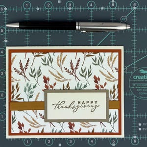 May include: A rectangular greeting card featuring a white background with a pattern of brown, rust, and sage green leaves. A horizontal mustard yellow ribbon crosses the center, topped with a rectangular tag reading "HAPPY thanksgiving".