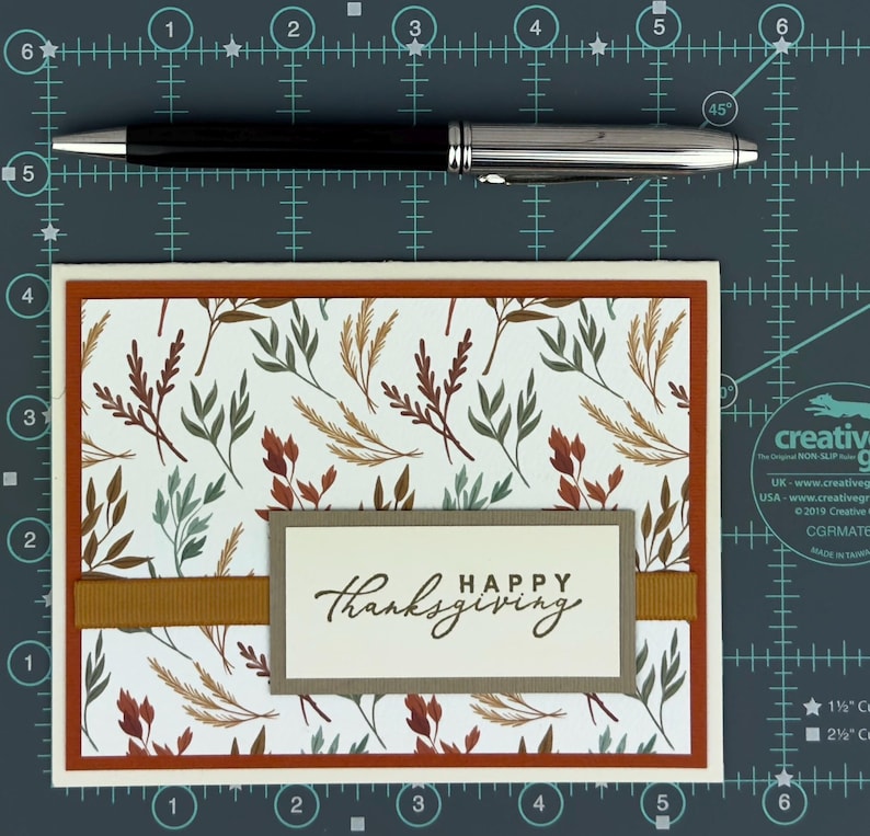May include: A rectangular greeting card featuring a white background with a pattern of brown, rust, and sage green leaves. A horizontal mustard yellow ribbon crosses the center, topped with a rectangular tag reading "HAPPY thanksgiving".