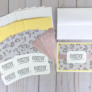 May include: A set of Easter card making supplies with a bunny pattern. The set includes card bases, envelopes, patterned paper, ribbon, and tags with the text "easter wishes".