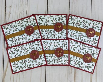 Può includere: Sei biglietti d'auguri rettangolari con sfondo bianco e un motivo ripetuto di foglie di agrifoglio verdi e bacche rosse. Ogni biglietto presenta una fascia orizzontale in nastro color senape e un sigillo in ceralacca rosso scuro con un fiocco dorato.